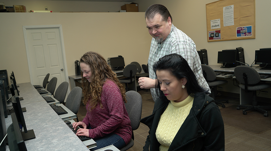 two students working in computer room, receiving assistance from instructor